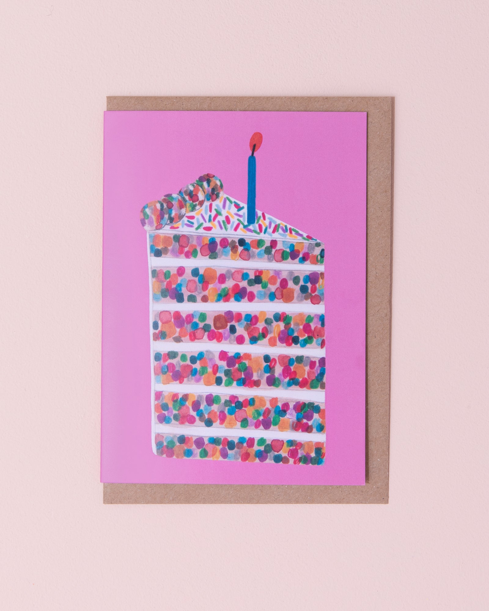 Daria Solak illustrated greetings card with brown kraft envelope. Slice of confetti birthday cake with a candle on top. Pink background