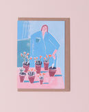 Luiza Holub illustration greetings card in blue, red and burgundy with a brown kraft envelope . Woman potting flowers and plants on a table.