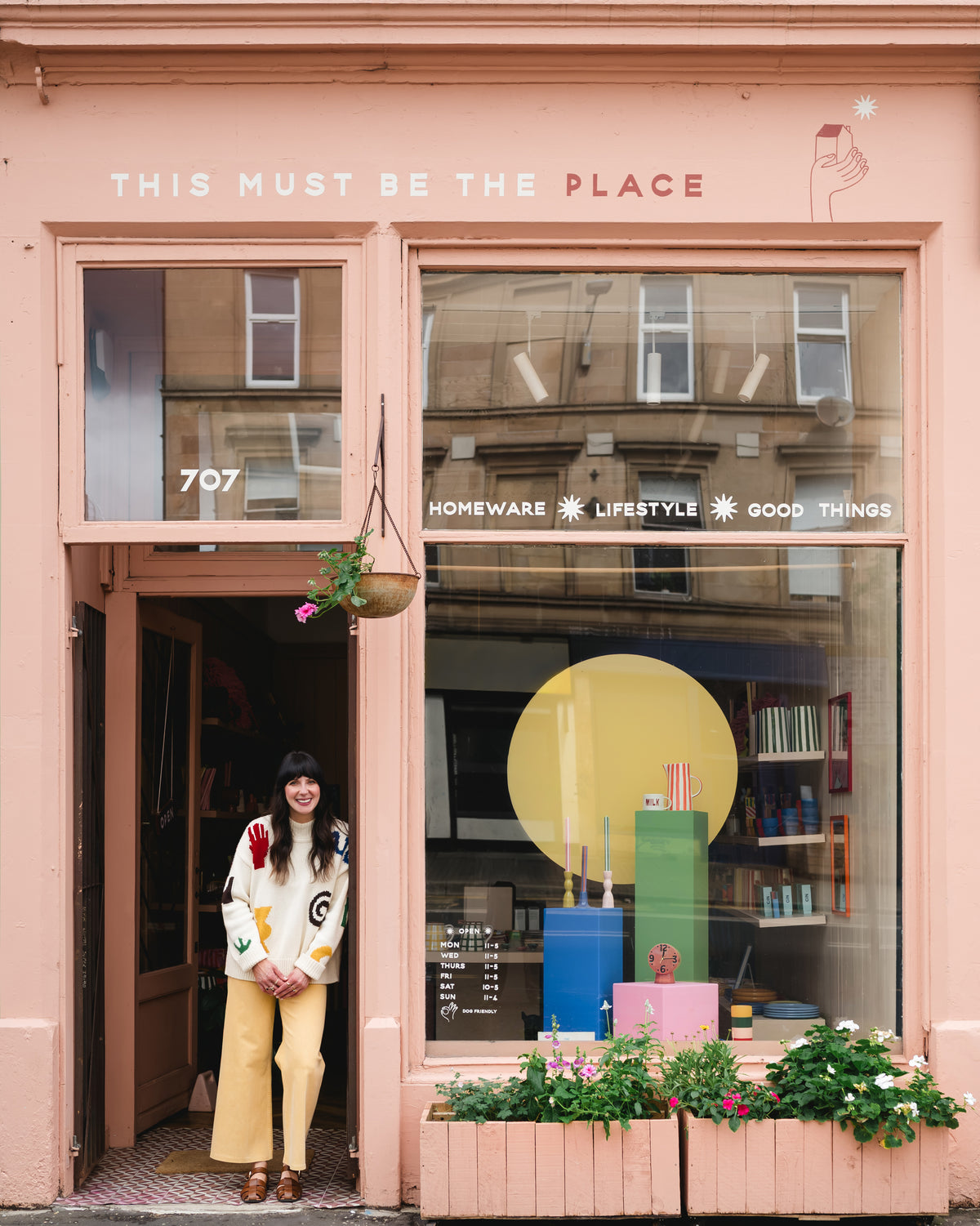 A woman stands in the doorway of a pink storefront with the words 'THIS MUST BE THE PLACE' above the entrance. The shop window displays colorful decor, including a large yellow sun graphic, books, and plants.